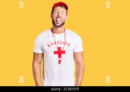 Young caucasian man wearing lifeguard t shirt holding whistle sticking tongue out happy with funny expression. emotion concept. Stock Photo