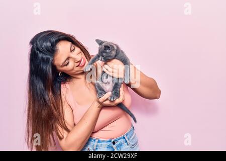 Young beautiful latin woman smilling happy. Standing with smile on face holding adorable cat over isolated pink background Stock Photo