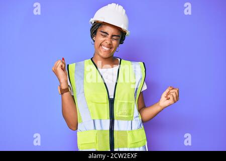 Young african american woman with braids wearing safety helmet and reflective jacket very happy and excited doing winner gesture with arms raised, smi Stock Photo
