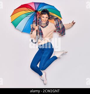 Young beautiful girl wearing winter clothes and scarf smiling happy. Jumping with smile on face holding colorful umbrella over isolated white backgrou Stock Photo