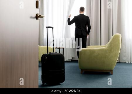Rear view of businessman wearing dark suit standing in hotel room looking into window Stock Photo