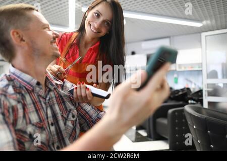 Woman hairdresser smiles and holds hair clipper in her hands and communicates with client. Stock Photo