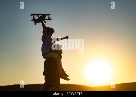 Sunset silhouette of Father carrying his son on shoulders. Boy child is sitting on daddy shoulder piggyback while the flight. Stock Photo
