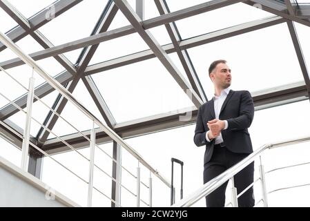 Young businessman wearing formal suit standing on a stairs in modern building with suitcase Stock Photo