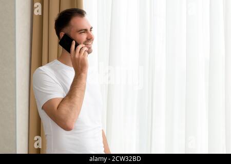 Handsome Caucasian man looking through window in hotel while speaking on smartphone Stock Photo