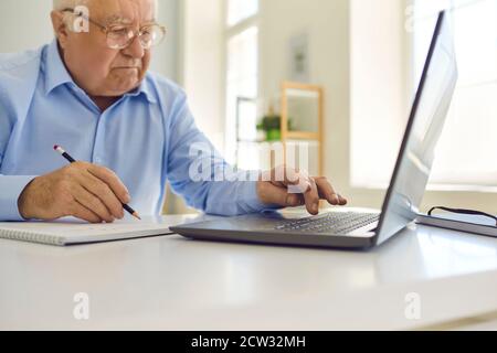 Senior man in glasses using laptop and taking notes in notebook sitting at desk at home Stock Photo