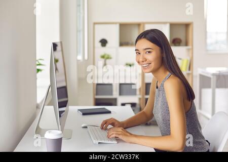 Young woman smiling and looking at camera while sitting at desk and working on computer