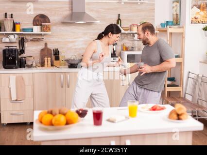 Couple having fun in the kitchen fencing with big spoons during breakfast wearing pajamas. Cheerful carefree joyful funny lovers, fighting wooden spoon, bonding game fight swordplay, happy lifestyle Stock Photo