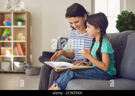 Happy mom and her daughter reading book of children's stories sitting on sofa in cozy room Stock Photo