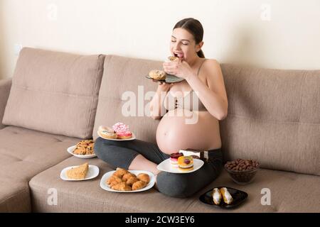 Hungry pregnant woman sitting on the sofa is eating a lot of unhealthy food such as donuts, cakes, chocolate cereal balls, croissants and cookies. Swe Stock Photo