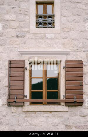 A brown window with wooden shutters and a small window with metal bars on a white wall. Stock Photo