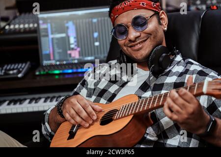 Close up portrait of happy young man, male artist in sunglasses smiling while playing ukulele, sitting in recording studio Stock Photo