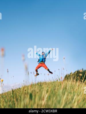 Backpacker traveler emotionally jumping over green grass mountain meadow with a backpack with wide opened arms and legs Stock Photo