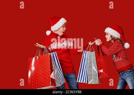 Cute little girl in Santa hat hanging shopping bag with Christmas gifts on hand of astonished boy during holiday celebration against red background. Stock Photo