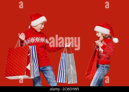 Cute little girl in Santa hat hanging shopping bag with Christmas gifts of astonished boy during holiday celebration against red background. Stock Photo