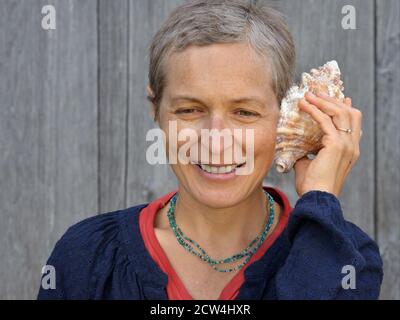 Modern middle-aged Caucasian Canadian country woman with short hair listens to a sea shell. Stock Photo