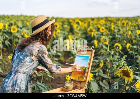 A young woman with curly hair and wearing a hat is painting in nature. A woman stands in a sunflower field on a beautiful day. Stock Photo