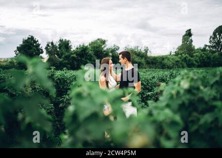 Young loving couple, woman and man, gently hugging with eyes closed on the background of green currant field. Foreground with blurred leaves. Stock Photo
