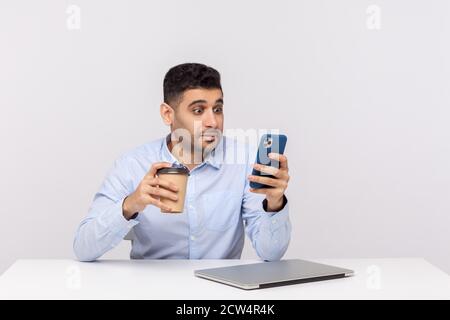 Amazed shocked businessman sitting at workplace with coffee in hand and checking message, reading email on mobile phone with surprised expression. ind Stock Photo