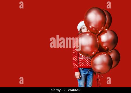 Cute child in red Santa hat and sweater hiding behind balloons on red background. Stock Photo