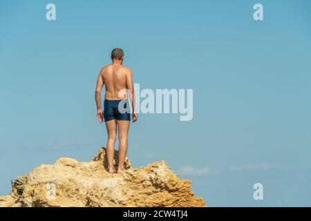 man in blue swimming trunks stands on top of a cliff against a blue sky. photo from the back Stock Photo