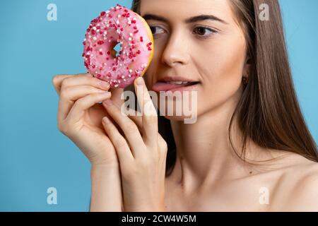 Woman holding a donut and sticking out tongue. Female model a donut in front of her face on blue background. Stock Photo