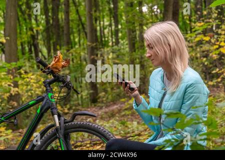 blonde girl in a turquoise jacket sits on a fallen tree in the autumn forest and looks at the phone and smiles, there is a Bicycle next to her Stock Photo