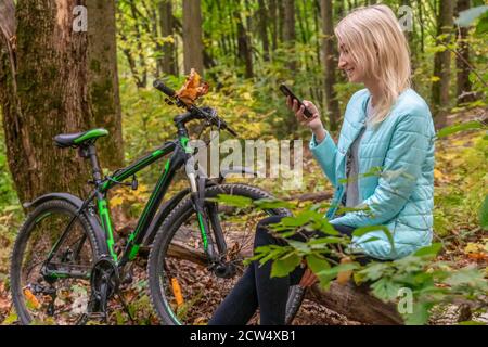 blonde girl in a turquoise jacket sits on a fallen tree in the autumn forest and looks at the phone and smiles, there is a Bicycle next to her Stock Photo