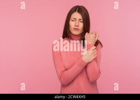 Sad brunette woman grimacing feeling pain in wrist, risk of bone fracture, dislocation, rheumatoid arthritis. Indoor studio shot isolated on pink back Stock Photo