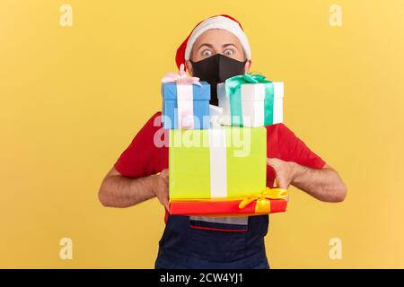 Male courier in red t-shirt, overalls and santa claus hat, with protective black mask on face delivering presents and gifts during coronavirus. Indoor Stock Photo
