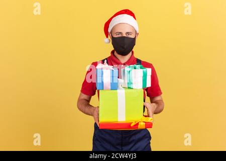 Deliveryman in black protective mask and santa claus hat standing in uniform and holding parcels in hands, delivery service during coronavirus. Indoor Stock Photo