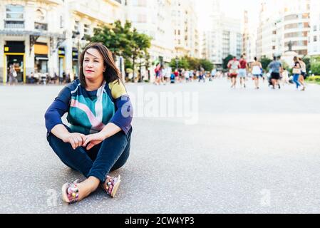 brunette woman in casual clothes sitting on the ground in a square in Europe with people, looking into the distance. copy space Stock Photo