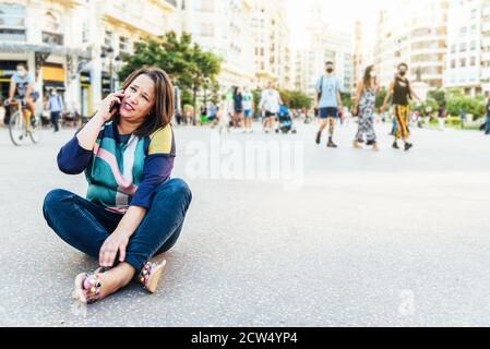brunette woman in casual clothes sitting on the ground in a square in Europe with people, talking on mobile phone. copy space Stock Photo