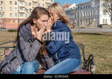 Portrait of a girl telling secrets to her astonished friend sitting on a park bench Stock Photo