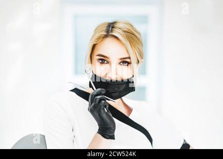 Portrait of young blond female dentist holding dental instruments. A woman wearing black mask and gloves. Space for text. Stock Photo
