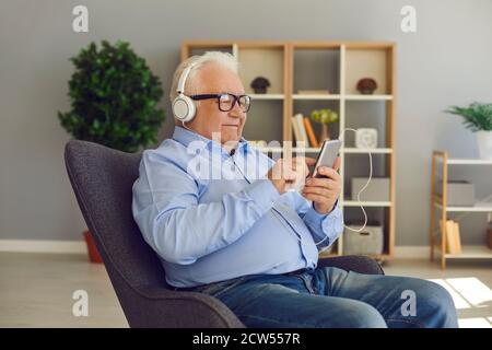 Happy senior man listening to favorite music or podcast wearing headphones and using smartphone Stock Photo