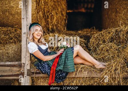 Cute blonde in dirndl, traditional festival dress with bouquet of field flowers sitting on the wooden fence at the farm with haystacks behind Stock Photo