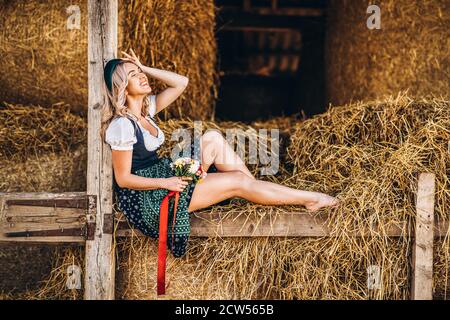 Cute blonde in dirndl, traditional festival dress with bouquet of field flowers sitting on the wooden fence at the farm with haystacks behind Stock Photo