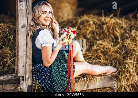 Cute blonde in dirndl, traditional festival dress with bouquet of field flowers sitting on the wooden fence at the farm with haystacks behind Stock Photo