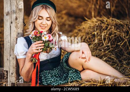 Cute blonde in dirndl, traditional festival dress with bouquet of field flowers sitting on the wooden fence at the farm with haystacks behind Stock Photo