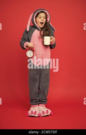 Drinking coffee energy. Morning routines. Adorable bunny hold alarm clock. Small girl in bunny costume. Child rabbit kigurumi. Girl bunny pajamas. Bunny kid red background. Baby animal. Counting time. Stock Photo