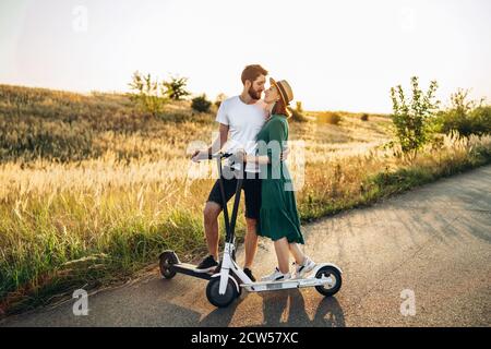 Portrait of young couple at sunset with beautiful natural landscape. Walking on electric scooters in the countryside. Stock Photo
