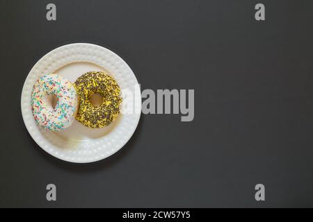 Glazed vanila and banana donuts with sprinkles on a plate on black stone background. Minimal flat lay with copy space Stock Photo