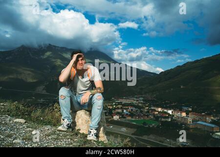 Young man grimaces, sitting on stone on background of village in mountains. Tired male tourist aping, resting on hill after active trekking in Stock Photo