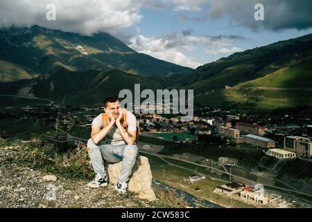 Young man grimaces, sitting on stone on background of village in mountains. Tired male tourist aping, resting on hill after active trekking in Stock Photo