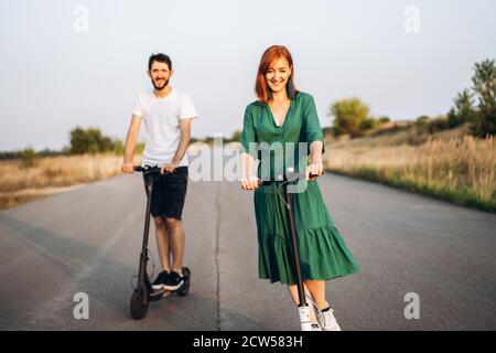 Young happy smiling couple having fun driving electric scooter on the road in the countryside. Content technologies. Stock Photo