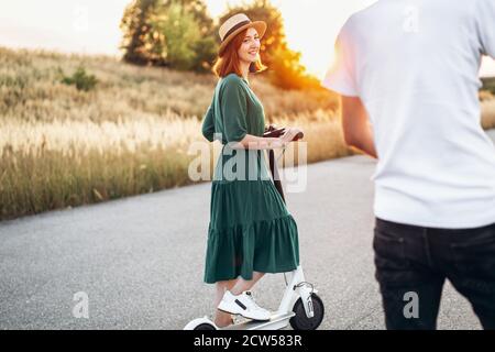 Happy couple walking on scooters. A young woman in dress and hat on sunset background. In the foreground, a man turns his back on the lens. Stock Photo