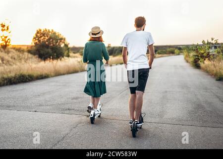 Happy couple walking on scooters. A young woman in dress and hat on sunset background. In the foreground, a man turns his back on the lens. Stock Photo