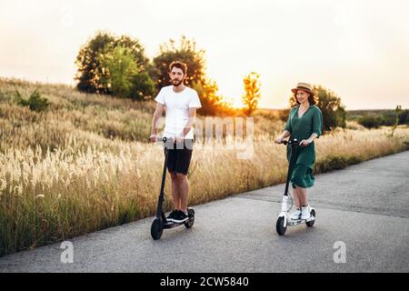 Happy couple walking on scooters. A young woman in dress and hat on sunset background. In the foreground, a man turns his back on the lens. Stock Photo