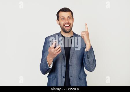 Young businessman man with a beard in a jacket using a smartphone surprised with an idea or question, pointing with a finger with a happy face, number one. Portrait of a man on a gray background. Stock Photo